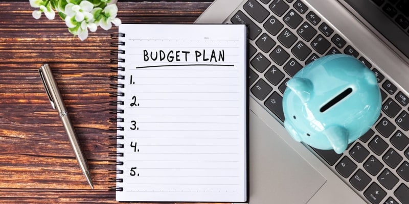 A flat lay of a budget planning workspace featuring a spiral notebook, silver pen, blue piggy bank, and laptop on a wooden surface.