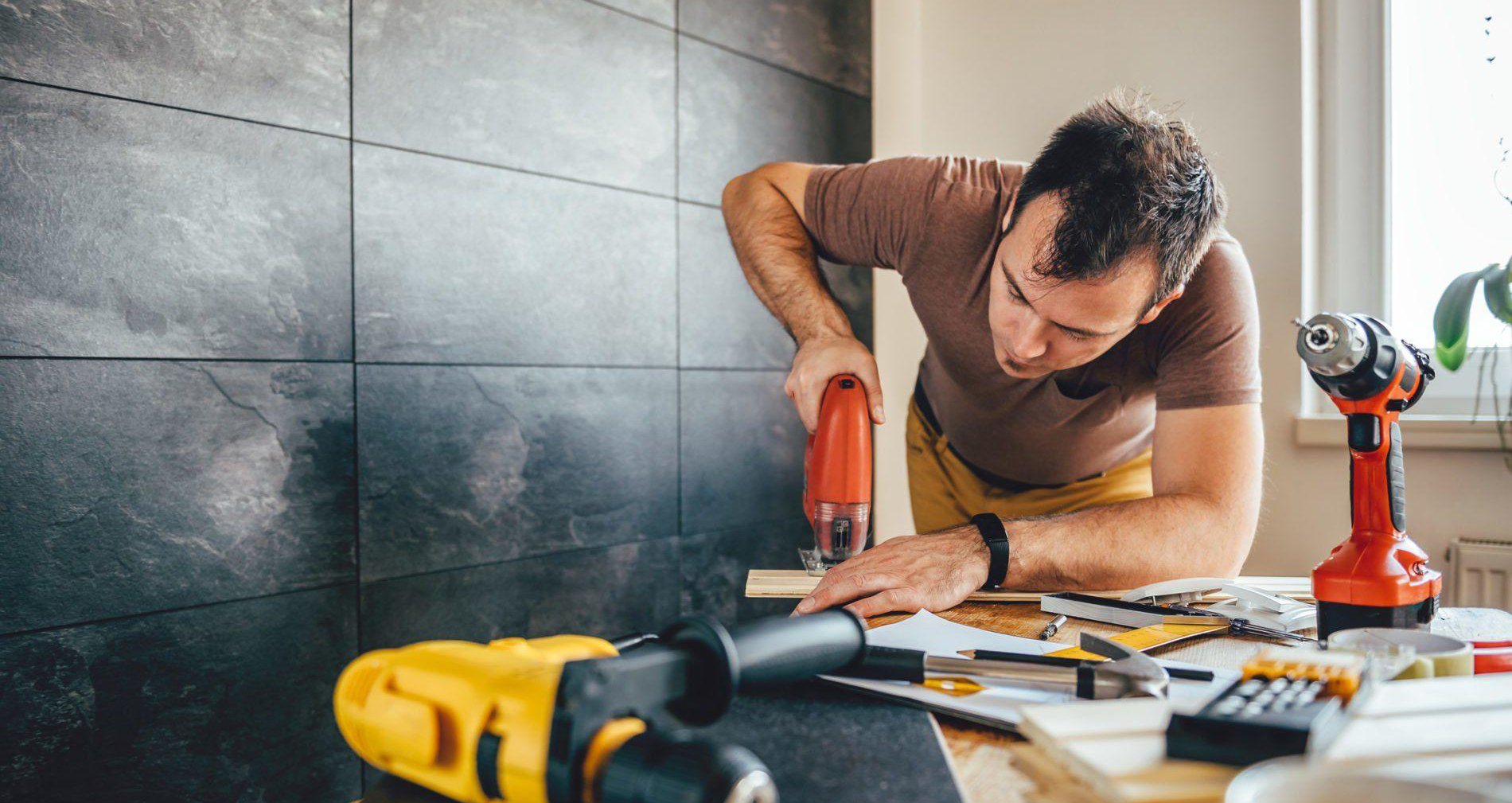 A person using a red jigsaw to cut wood on a cluttered workbench, surrounded by drills, tape measure, calculator, and renovation tools, with natural light from a nearby window.