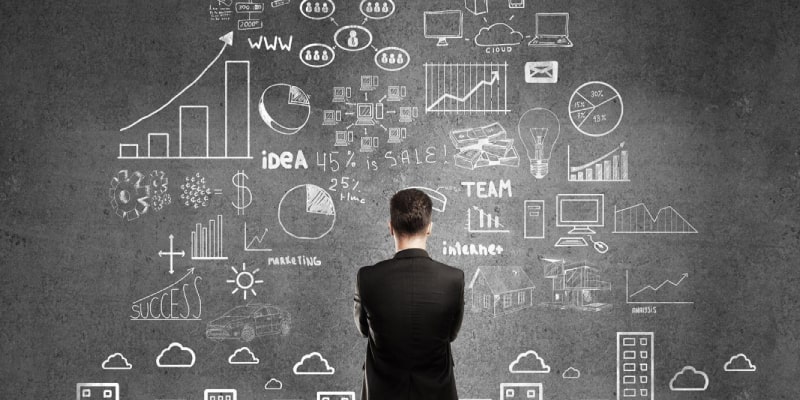 A person in a business suit stands before a dark wall filled with white chalk-style diagrams, graphs, and keywords like “IDEA,” “TEAM,” and “MARKETING,” symbolizing strategic planning and data analysis for renovation project timeline Dubai.