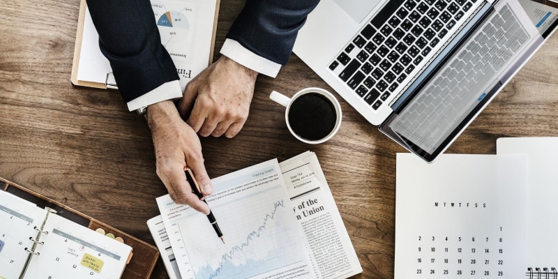 A professional workspace showing a person in a suit pointing at a rising financial graph, with a laptop displaying a spreadsheet, coffee cup, planner, and newspaper. The setup reflects strategic planning and financial analysis relevant to renovation project timeline Dubai.