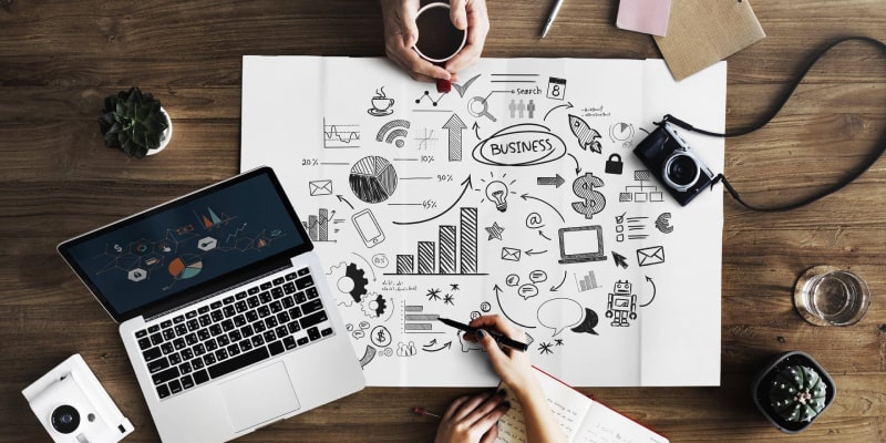 A collaborative workspace showing two people planning a renovation project. The table features hand-drawn business diagrams, a laptop with data charts, coffee, and mobile devices—symbolizing teamwork and strategic planning for renovation project timeline Dubai.