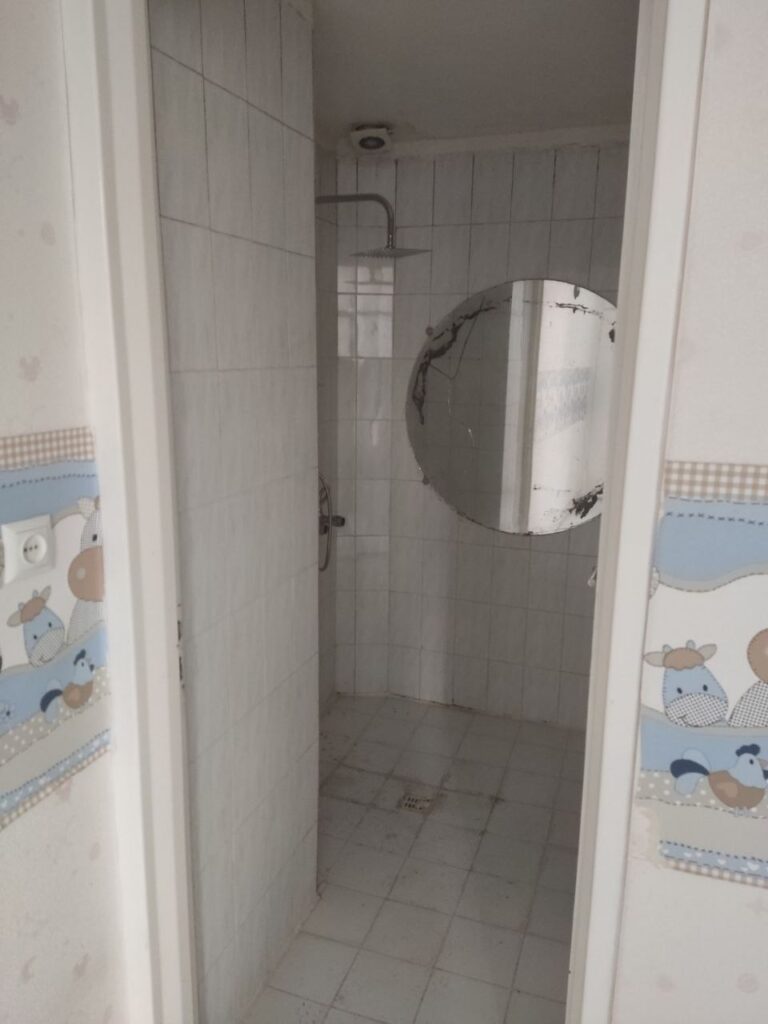An old outdated bathroom with a damaged round mirror and old white tiles before a complete modern renovation by Fardo Technical Services.
