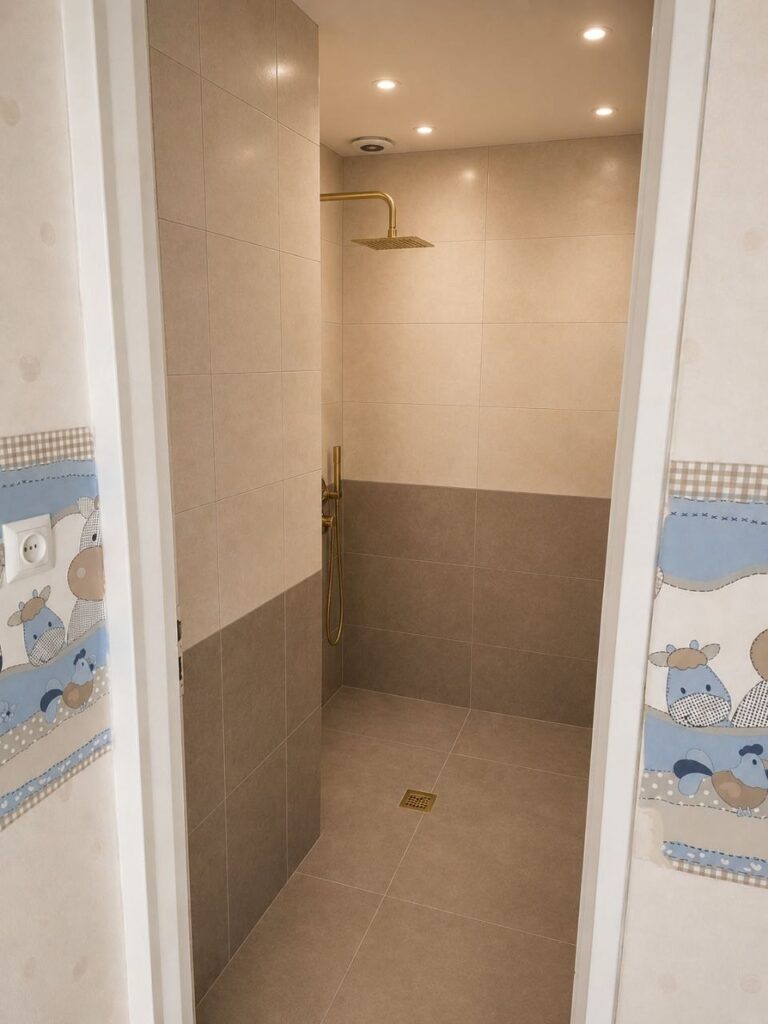 A modern renovated bathroom by Fardo Technical Services featuring two-tone beige and brown wall tiles, a luxury gold rain shower, and a matching gold floor drain.