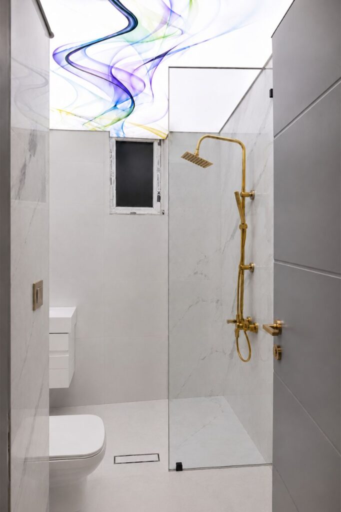 An elegant bathroom with a colorful printed abstract stretch ceiling, white marble-patterned tiles, and a gold rain shower set, designed by Fardo Technical Services.
