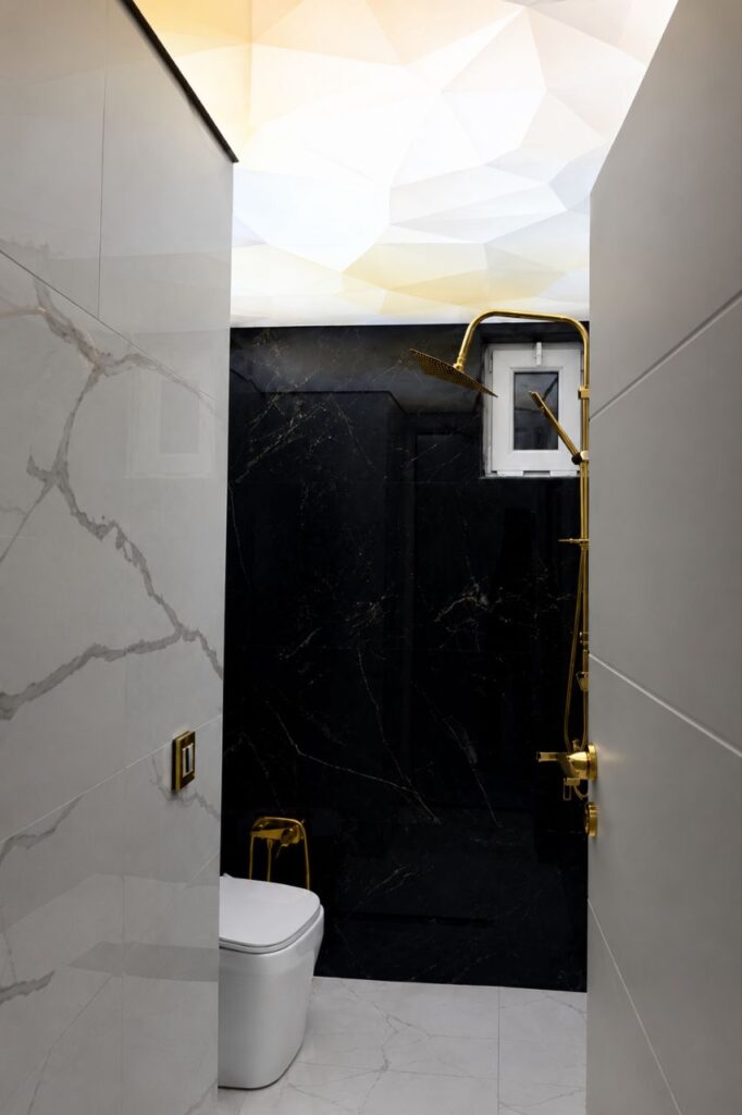 Bathroom entrance view by Fardo Technical Services highlighting a backlit geometric stretch ceiling, contrasting black marble walls, and a premium gold shower.